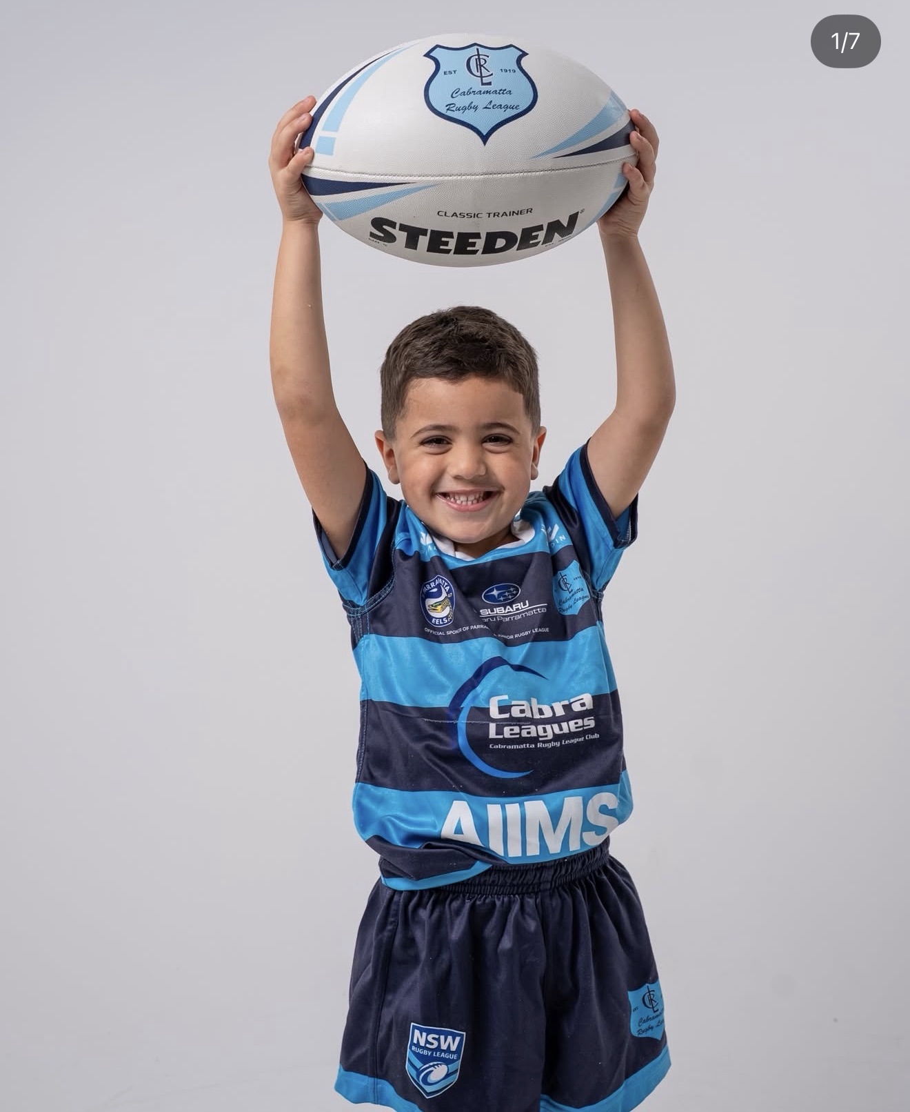 Junior Cabramatta Two Blues player holding a rugby league ball above his head in an AIIMS branded jersey